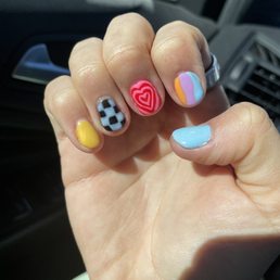 Nails done by Trinity - A person's hand with decorated nails featuring various colorful designs, including hearts, checks, and abstract patterns, is shown inside a vehicle.