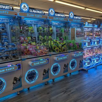 Claw Party Arcade - A row of claw machines filled with plush toys is shown in an arcade at Claw Party in Glendale, AZ. The machines are illuminated with blue neon lights, and each has signage reading 'Claw Party' and 'Prize Out!'. Plush toys of various characters and colors are visible inside the machines. The arcade has a wooden floor and a ceiling with fluorescent lighting. A large stuffed animal is placed on top of one of the machines. - Arcades Near Me - Glendale, Arizona