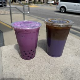 Ube latte and boba - Two iced beverages are positioned on a concrete surface outdoors. The drink on the left is purple with black tapioca pearls at the bottom, likely a taro or ube flavored drink. The drink on the right has a layered appearance with brown and purple hues, possibly an iced coffee or tea with flavoring. The background shows a street scene with cars, a sidewalk, and storefronts, indicating a casual outdoor setting near a coffee shop or cafe.