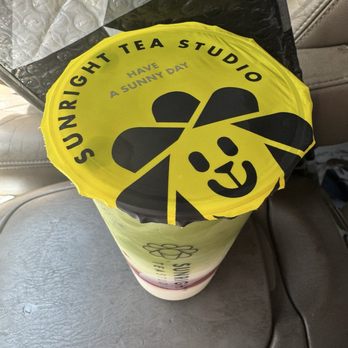 Sunright Tea Studio - Strawberry Matcha Latte with pudding - A beverage cup is covered with a yellow paper lid that has a panda face design and the text 'Sunright Tea Studio' and 'Have a Sunny Day.' The cup is positioned on a gray car seat, with some bubble wrap and packaging material nearby. The cup appears to contain a layered drink, possibly tea or milk tea, with a straw hole in the lid. - Bubble Tea Near Me - Fullerton, California