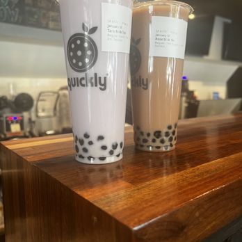 Quickly - Ann Arbor - Milk teas - Two large bubble tea drinks with tapioca pearls are positioned on a polished wooden counter at Quickly. One drink is white milk tea, and the other is brown milk tea, both in clear cups with lids. The cups feature the Quickly logo and labels indicating the drink types. The background shows a blurred cafe interior with equipment and a chalkboard menu. - Bubble Tea Near Me - Ann Arbor, Michigan