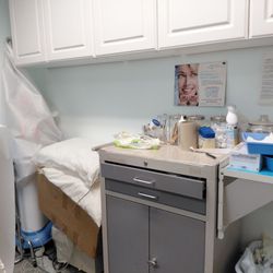 Micki Ly, MD - Piles of pillows next to table with a mess of medical supplies in the exam room. Vaseline not covered properly. Table cluttered and dirty