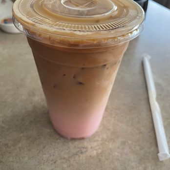 Cafe Fresco - A clear plastic cup contains iced coffee with visible ice cubes and a layered appearance, topped with a sealed plastic lid. The cup is on a beige table, with a straw wrapper lying next to it. The background includes a microwave and other kitchen items, indicating a casual dining or break area. - Cafes Near Me - Crown Point, Indiana