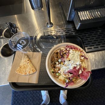 CAVA - my bowl with a pita bread - A tray holds a salad with cheese and vegetables, a slice of pita bread, and a glass of water on a counter at a restaurant or cafe. - Mediterranean Near Me - Cedar Park, Texas