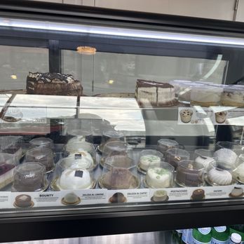 Cafe Select - More cakes - The image shows a glass display case containing multiple cakes and desserts, including chocolate, vanilla, and layered varieties. The cakes are arranged on different levels within the case, with some in plastic containers and others directly on the shelves. The reflection of the window and ceiling lights can be seen on the glass surface. The setting appears to be a bakery or cafe, with a focus on imported Italian cakes and desserts. - Coffee & Tea Near Me - Fort Lee, New Jersey
