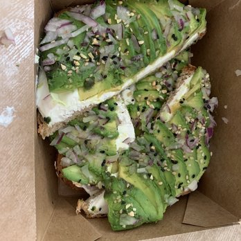 Compton's - Avocado Toast - Three slices of avocado toast topped with sliced avocado, chopped red onions, sesame seeds, and seasonings are arranged in a brown takeout box. The toast appears to be made with whole grain bread, and the toppings are evenly distributed across the slices. The image shows a close-up view of the food, highlighting the fresh ingredients and garnishes. - Sandwiches Near Me - Astoria, New York
