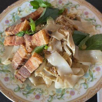 Glai Baan - Pad kee mao- Chinese broccoli, chili, garlic, Thai basil, mushroom, bamboo shoot, egg, rice noodle with crispy pork belly - A dish featuring crispy pork belly slices and rice noodles with vegetables, garnished with fresh basil and herbs. - Thai Near Me - Phoenix, Arizona
