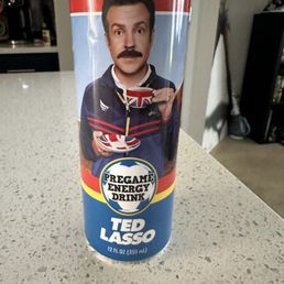 Energy Drink - The image shows a 12 oz (355 ml) energy drink can with a colorful label. The label features a man with dark hair and a mustache, holding a cup and a spoon, dressed in a jacket with red and blue stripes. The can's design includes the text 'Pregame Energy Drink' and 'Ted Lasso,' referencing the character from the television series. The can is placed on a speckled countertop in an indoor setting.