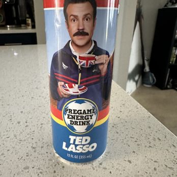 Atown - Energy Drink - The image shows a 12 oz (355 ml) energy drink can with a colorful label. The label features a man with dark hair and a mustache, holding a cup and a spoon, dressed in a jacket with red and blue stripes. The can's design includes the text 'Pregame Energy Drink' and 'Ted Lasso,' referencing the character from the television series. The can is placed on a speckled countertop in an indoor setting. - Accessories Near Me - Austin, Texas