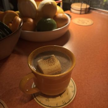 Double Fun - A ceramic mug filled with hot chocolate topped with a large marshmallow is placed on a coaster on a restaurant table. In the background, there are bowls of fruit, including lemons, limes, and possibly oranges, on the table. The setting appears to be indoors with warm lighting, and the table surface is a reddish color. The scene suggests a cozy dining or dessert experience. - Cocktail Bars Near Me - Chicago, Illinois