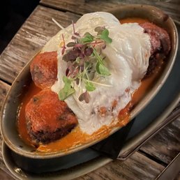 Eggplant Meatballs - The image shows a ceramic dish containing eggplant meatballs topped with melted cheese and garnished with microgreens. The dish is placed on a rustic wooden table, with a bowl and utensils visible nearby. The presentation emphasizes a hearty, vegetarian-friendly meal with fresh garnishes and a flavorful sauce.