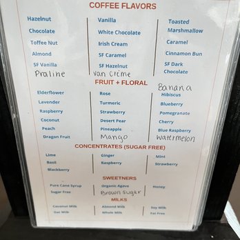Cafe Bebida - The menu board features sections for coffee flavors, including hazelnut, vanilla, and toasted marshmallow, with handwritten notes like 'Praline' and 'van Crème.' It also offers fruit and floral options such as banana, mango, and blueberry, along with sugar-free concentrates like lime and ginger. Sweeteners include organic agave and honey, and milk options include coconut, almond, and soy milk. The board is framed with a black border and is placed on a surface, likely at a café. - Coffee & Tea Near Me - Goodyear, Arizona