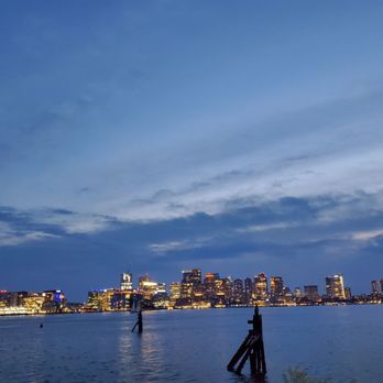 Hyatt Regency Boston Harbor - Gorgeous view! - The image depicts a city skyline at dusk, with numerous buildings lit up along the waterfront. The sky is partly cloudy with shades of blue, and the water in the foreground reflects the city lights. There are some wooden posts in the water, indicating a harbor or dock area. The scene captures an urban landscape with a calm water body and a vibrant cityscape. - Hotels Near Me - Boston, Massachusetts