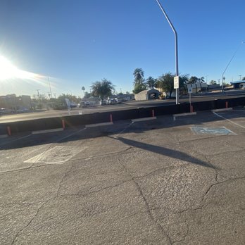 The Drink All - The image shows an empty parking lot with visible cracks on the asphalt surface. There is a designated handicap parking space marked with blue paint and a wheelchair symbol. A raised curb runs across the lot, with red posts along its edge. The background features trees, buildings, and a clear blue sky, with the sun shining brightly from the left side of the image, creating glare and shadows. - Beer, Wine & Spirits Near Me - Mesa, Arizona