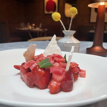 Lyle - STRAWBERRIES Rhubarb, Lavender, Lime - A plated dessert featuring strawberries and rhubarb, garnished with a mint leaf and decorative elements, set on a table with a vase and lamp. - Hotels Near Me - Washington, DC, District of Columbia