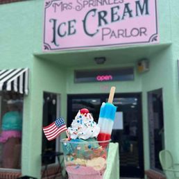 A cup of ice cream topped with whipped cream, sprinkles, a cherry, and an American flag, held outside Mrs. Sprinkles Ice Cream Parlor.