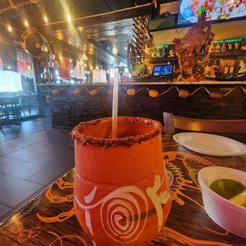 Mariscos El Puerto 2 - A beverage served in a glass with a salted rim and a straw, placed on a wooden table in a restaurant setting. The background features a bar area with bottles, a mounted fish sculpture, and a television screen displaying food. The restaurant has warm lighting and decorative string lights. - Seafood Near Me - Chicago, Illinois