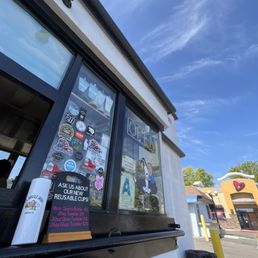 Outside window - A coffee shop window displays multiple stickers and signs, including an 'OPEN' sign and a cartoon character holding a coffee cup. The window has a black frame, and there is a small sign advertising reusable cups and drink sizes. The sky above is clear with some wispy clouds, and part of the building exterior, including a trash can and a yellow post, is visible outside.