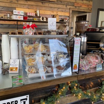 Blackjack’s - A coffee shop counter features a glass display case filled with muffins and baked goods, each in paper liners. Behind the counter, a wooden wall with a Blackjack's sign is visible, along with shelves holding various items. To the right, a coffee grinder and jars of candies are present, with a small sign listing flavors. The counter is decorated with a garland of greenery and lights, creating a festive atmosphere. - Coffee & Tea Near Me - Dublin, Texas