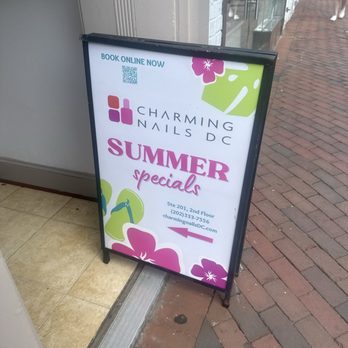 Charming Nails - A sidewalk A-frame signboard displays promotional information for Charming Nails DC, a nail salon. The sign features the salon's logo, name, and details about summer specials. It includes the address, phone number, website, and an arrow pointing to the entrance. The sign is decorated with floral graphics in pink, green, and purple, and is positioned on a brick sidewalk outside the salon, which has large glass windows and a white brick exterior. - Nail Salons Near Me - Washington, DC, District of Columbia