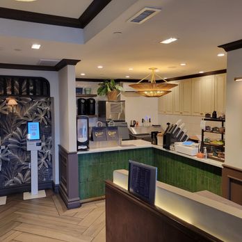 Caphe Den - inside - The photo depicts the interior of a coffee shop featuring a counter area with a digital menu display on the wall showing various drink options. Two digital ordering kiosks are positioned to the left, and the counter has equipment and supplies for beverage preparation. The decor includes a chandelier, cabinetry, and decorative elements, with a clean and organized layout. - Coffee & Tea Near Me - Flushing, New York
