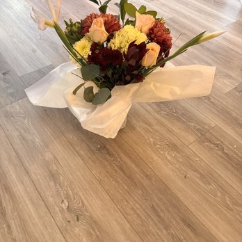 Produce Junction - She was thrilled with her bouquet! - A floral arrangement with roses, lilies, and other flowers wrapped in white paper, placed on a wooden floor. - Fruits & Veggies Near Me - Exton, Pennsylvania