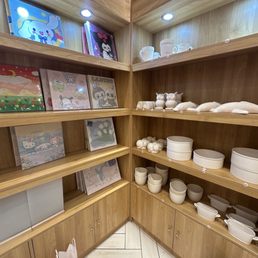 Wooden shelves in a retail or display area contain colorful illustrated artwork, including cartoon characters and scenic designs, on the upper shelves. The middle shelves feature white ceramic mugs, animal figurines, and various white ceramic containers and bowls. Spotlights installed in the ceiling highlight the display, and the lower cabinets are closed with small round knobs. The setting appears to be a shop or showroom for ceramic or decorative items.