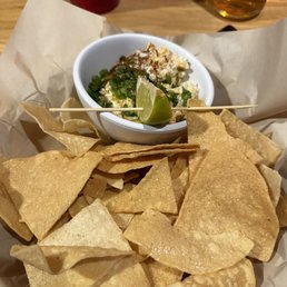 Elote dip - a must get - A serving of crispy tortilla chips surrounds a small white bowl of dip topped with chopped green onions and cheese. A lime wedge is placed on the edge of the bowl. The chips are arranged on parchment paper on a wooden table, with drinks visible in the background. The setting appears to be a casual dining environment, possibly a restaurant or bar.