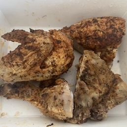 Portion given for 1/2 Chicken! - Six pieces of grilled chicken, including drumsticks and thighs, are placed inside a white takeout box. The chicken has visible seasoning and grill marks, indicating it was cooked on a grill or open flame. The container shows some juices and seasoning residue.