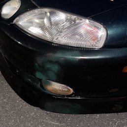 Al smashed up my son's car and then attempted to hide the $800 in damage with spray paint. - A close-up view of the front left side of a dark-colored vehicle, highlighting the headlight, bumper, and part of the hood. The car has some scratches, dirt, and minor damage on the bumper area. The vehicle is parked on a paved surface with some small debris and leaves nearby.