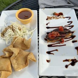 Lobster crab Rangoon and steak bruschetta - Two rectangular white plates with food are placed on a black outdoor metal table. The left plate contains fried appetizers, coleslaw, and a small bowl of dark sauce. The right plate features a cooked steak topped with cherry tomatoes, drizzled with balsamic glaze, and garnished with capers. Silverware and a gray napkin are also visible on the table, with a grassy area in the background.