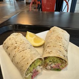 Falafel Wrap - A white square plate holds two falafel wraps, each filled with chopped vegetables and falafel, wrapped in grilled flatbread. A lemon wedge is placed beside the wraps. The background shows a restaurant interior with tables, chairs, and large windows, indicating a casual dining environment.