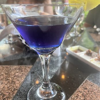 Desert Bistro - Violet blues - A blue-colored cocktail served in a martini glass on a restaurant table, with a lemon garnish and various dishes and drinks in the background. - New American Near Me - Moab, Utah