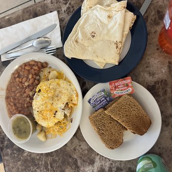 Ramblin'cafe - two plates of food on a table - The image shows a breakfast setup on a marble table, featuring a plate with beans, scrambled eggs topped with cheese, and a small container of green sauce. There is a separate plate with folded tortillas, a plate with two slices of toasted bread, and small containers of jelly and jam. Beverages include a bottle of orange-colored drink and a can of soda. Utensils and a napkin are also visible on the table. - Kosher Near Me - Santa Fe, New Mexico
