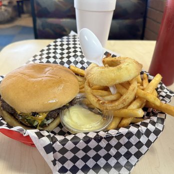 Rex's Hamburgers - Yum - A meal featuring a cheeseburger with a beef patty, lettuce, and cheese in a bun, accompanied by a serving of French fries, onion rings with a plastic spoon inserted, and a small container of butter or margarine. The food is presented in a checkered paper-lined basket on a light-colored table, with a white cup and a red ketchup bottle visible in the background. - Burgers Near Me - Albuquerque, New Mexico