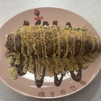 Chocolate Bash - Bash Roll - A dessert, possibly a banana or similar item, is placed on a pink plate and covered with drizzles of chocolate and green sauce. It is topped with shredded toppings, which could be chocolate, nuts, or similar ingredients. The plate has a strawberry logo and some icons around the edge, indicating it is from a dessert or snack business. - Desserts Near Me - Manteca, California