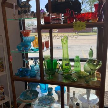 Blessed Again - The image shows a wooden and glass display shelf filled with an assortment of colorful glassware, including vases, bowls, and cups, arranged on multiple shelves. The shelf is placed near a window, allowing natural light to illuminate the items and reveal outdoor scenery with trees and a street. Some items are green, blue, red, and clear, with different shapes and designs, and a black bag is visible on top of the shelf. - Used, Vintage & Consignment Near Me - Granbury, Texas