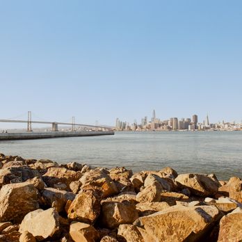 Photolab - The image depicts a view of the San Francisco skyline across a body of water, with numerous high-rise buildings visible in the distance. In the foreground, there are large rocks along the shoreline, and a pier extends into the water on the left side of the image. The sky is clear and blue, indicating a sunny day. - Photography Stores & Services Near Me - Berkeley, California