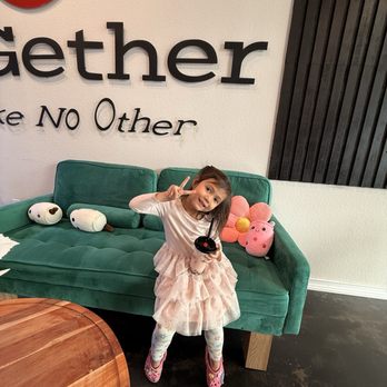 Teagether - Strawberry milk tea boba - A young girl with dark hair, wearing a pink dress with a ruffled skirt and patterned leggings, sits on a green sofa inside a cafe. She holds a drink with a straw and makes a peace sign with her right hand. Plush toys, including a white dog and a pink pig, are placed on the sofa behind her. The cafe has a white wall with black lettering and a black wooden panel on the right side. - Bubble Tea Near Me - Grand Prairie, Texas