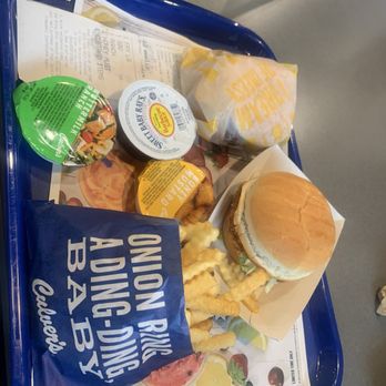 Culver's - Spicy chicken sandwich - A blue plastic tray contains a burger with a bun, lettuce, and other fillings, along with a serving of crinkle-cut fries. There are small containers of salad dressing and a cup of sauce, with a wrapped sandwich or burger in the background. The tray is placed on a table with a paper menu underneath, and part of a person's hand is visible at the top of the image. - Burgers Near Me - St. Cloud, Florida