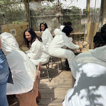 Choplet - Several women dressed in white protective suits are seated on chairs and a wooden bench inside a greenhouse or craft studio with transparent plastic walls and a wooden floor. They appear to be participating in a pottery or craft workshop, with some working on projects and others observing. The space is decorated with string lights and has a natural, plant-filled background visible through the transparent walls. - Art Schools Near Me - Brooklyn, New York