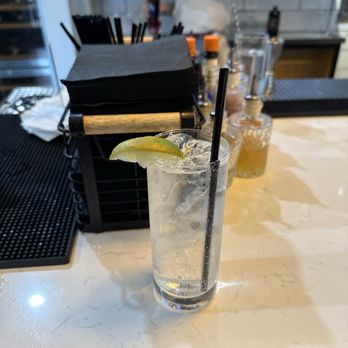 Penelope - Tito's Soda - A tall, clear glass filled with Tito's Soda, ice cubes, and a lemon wedge on the rim, is positioned on a bar counter. The background includes bar tools, bottles, and a person standing behind the bar, partially visible. The setting appears to be a bar or restaurant with a casual atmosphere. - Pizza Near Me - Tucson, Arizona