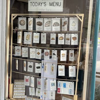 Breadboard Bakery - A bakery storefront window features a menu board with images and prices of baked goods, pastries, and drinks, including sourdough, bread, and tea. - Bakeries Near Me - Arlington, Massachusetts