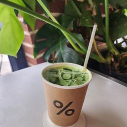 Matcha latte - A paper cup containing a green matcha latte with ice, placed on a white table near green plants and a brick wall.