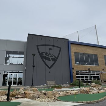 Topgolf - Curb appeal - The image shows the exterior of a modern building with a large logo on the wall, adjacent to a mini golf course with artificial turf and rocks. - Sports Bars Near Me - Mobile, Alabama