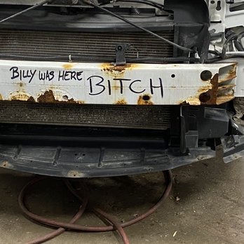 North East Auto Body - This is what they put on my wife's car when they did all the work an it was never there before an that's how - The image depicts a damaged car bumper with rust spots and corrosion, attached to a vehicle in a repair shop. Handwritten text on the bumper reads 'Billy was here Bitch.' The background includes visible hoses and a concrete floor, indicating an automotive repair setting. The bumper appears to be removed or in the process of repair, with signs of wear and damage. - Body Shops Near Me - Johnston, Rhode Island