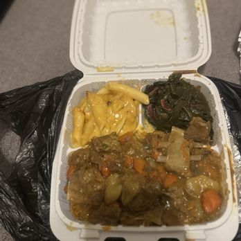 Traphouse Caribbean - Curry Goat Mac N Cheese Greens - A plastic takeout container holds macaroni and cheese, cooked greens, and beef stew with carrots and potatoes, placed on a gray surface. - Caribbean Near Me - Austin, Texas