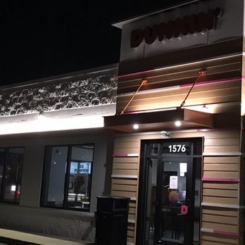 Dunkin' - The image shows the exterior of a Dunkin' store at night, with illuminated signage displaying the Dunkin' name. The building has a contemporary design with decorative lighting along the upper facade and a combination of wood and metal exterior materials. Large windows reveal the interior, and the entrance is centered with a glass door. The address 1576 is visible above the door, and the scene is set against a dark night sky. - Breakfast & Brunch Near Me - Columbus, Ohio