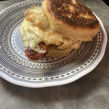 Rehoboth Baking - Sausage, egg, and cheese on a grilled English muffin - A breakfast sandwich on a decorative black and white patterned plate includes a toasted English muffin with a browned top, a fried egg with slightly runny yolk, a sausage patty, and melted cheese. The sandwich is placed on a metal table surface, with a blurred background. The image highlights a hearty breakfast item typically served at a bakery or cafe. - Bakeries Near Me - Rehoboth, Massachusetts