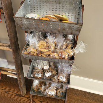 Desserts Etc  -  display of pastries and pastries - A four-tier metal display stand showcases an assortment of baked goods, including cookies and muffins, some in clear plastic bags with labels. The stand is positioned on a wooden floor against a neutral-colored wall, indicating an indoor bakery or coffee shop environment. The top tier has unwrapped baked items, while the lower tiers contain packaged treats, some labeled gluten-free. - Desserts Near Me - Hershey, Pennsylvania